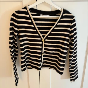 Randig kofta från mango med guldknappar - Snygg svart och vit randig kofta från mango i storlek XS. Koftan har v-ringning, guldknappar framtill och på axlarna samt långa ärmar. Perfekt att slänga över en topp för en cool och klassisk look.