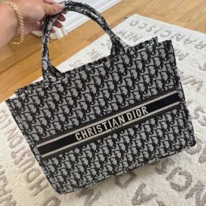 Christian Dior Book Tote svart/vit - Säljer en ikonisk Christian Dior Book Tote i svart och vitt med det klassiska Dior-mönstret. Väskan är rymlig med två handtag och har broderad logga framtill. Perfekt för dig som vill ha en statementväska med lyxig känsla och snygg design.