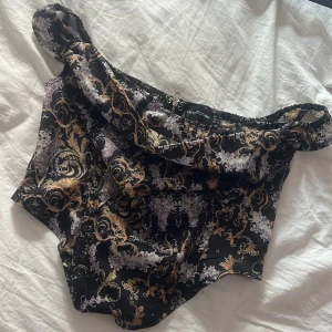 Svart offshoulder topp PrettyLittleThing - Säljer en svart offshoulder topp från PrettyLittleThing med barockinspirerat mönster i guld och vitt. Toppen har en croppad passform och spetsig nederkant, storlek 36 