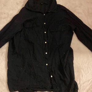 Svart skjorta från Miss Millo - Svart skjorta från Miss Millo i klassisk modell med långa ärmar och vita knappar framtill. Skjortan har två bröstfickor och en avslappnad passform. Perfekt att styla med jeans eller kjol för en cool look.