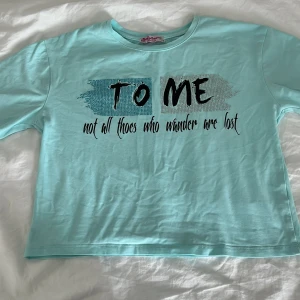 Turkos croppad t-shirt från Gjollen - Turkos croppad t-shirt från Gjollen med svart texttryck 'To me not all thoes who wonder are lost' framtill. Klassisk rund halsringning och korta ärmar. Skön och lätt i bomull, perfekt för en chill stil.