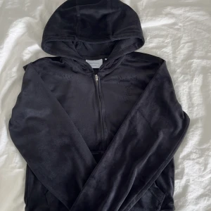 Svart hoodie från Juicy Couture - Svart hoodie från Juicy Couture i mjuk velour med dragkedja framtill och huva. Klassisk JC-logga broderad på bröstet och känguruficka. Perfekt för en chill och trendig look.