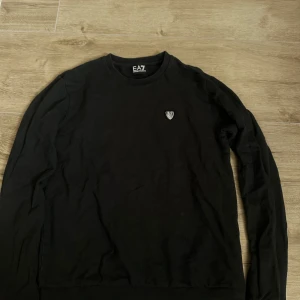 Svart EA7 crewneck tröja - Svart långärmad crewneck tröja från EA7 Emporio Armani. Klassisk och stilren design med EA7-logga på bröstet. Tillverkad i mjuk bomull som känns skön mot huden. Perfekt för dig som gillar en clean och enkel look.