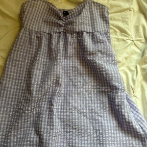 Ljuslila rutig axelbandslös klänning - Söt axelbandslös klänning i ljuslila och vitt rutigt mönster. Klänningen har en lätt v-ringning och är luftig med en avslappnad passform, perfekt för varma dagar. Materialet känns mjukt och bekvämt mot huden.