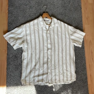 Randig kortärmad skjorta Jack & Jones - Snygg kortärmad skjorta från Jack & Jones i beige och vitt med tunna bruna och grå ränder. Skjortan har klassisk krage, knäppning framtill och avslappnad passform. Perfekt för varma dagar och enkel att styla med jeans eller shorts.