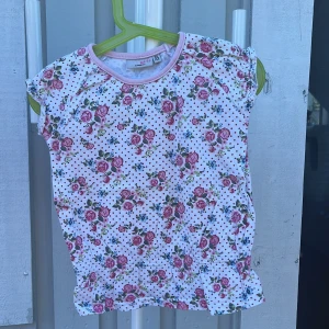 Blommig topp Mini Girls KappAhl - Supersöt topp från Mini Girls by KappAhl i storlek 98/104. Vit botten med små röda prickar och blommigt mönster i rosa, grönt och blått. Kortärmad modell med volangkant nertill och ljusrosa kant vid halsen. Perfekt för vår och sommar!