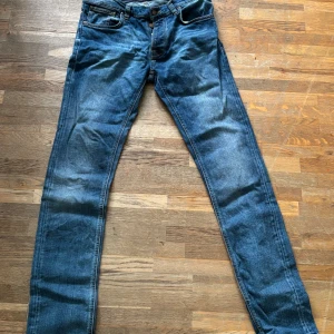 Blå jeans från Nudie Jeans - Säljer ett par klassiska blå jeans från Nudie Jeans med orange sömmar och snyggt broderad bakficka. Jeansen har normal passform och raka ben, tillverkade i slitstarkt bomullstyg. Perfekta för dig som gillar en tidlös denimlook. Inga hål eller skador på jeansen. Säljer där med att byxorna är lite för långa på mig.