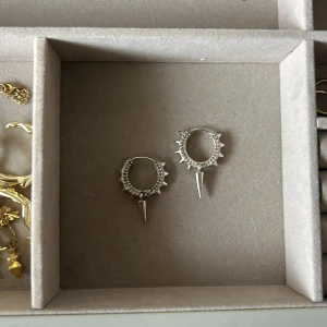 Silvriga örhängen med nitar och spikes - Coola silvriga örhängen i ringmodell med små nitar runt hela ringen och en spetsig spike som hänger ned från Syster P. Perfekta för dig som gillar edgy och unik stil. Passar till både streetwear och rockiga outfits.