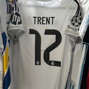 Real Madrid Trent 12 Champions League fotbollströja - Snygg vit Real Madrid fotbollströja från Adidas med svarta detaljer och gula inslag. Tryck med Trent och nummer 12 på ryggen, Champions League-märke på ärmen och klubbmärke på bröstet. Materialet är lätt och andas, perfekt för match eller träning.