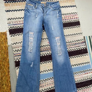 Slitna blå, lågmidjade bootcut jeans - Snygga ljusblå bootcut jeans från DDET Jeans med slitna detaljer och råa kanter. Jeansen har klassisk femficksdesign, låg midja och dragkedja. Innerbenslängd 80cm