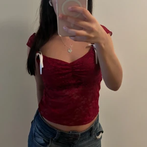 Vinröd spetsig croppad topp med rosetter - Säljer en vinröd croppad topp i spets med korta ärmar och vita rosetter på axlarna. Toppen har en lätt rynkad detalj vid bysten och är figurnära. Perfekt för dig som gillar romantiska och trendiga plagg med det lilla extra.