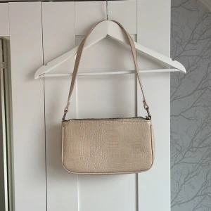 Beige axelväska med krokoprägling - Säljer en beige axelväska med krokodilpräglat mönster och dragkedja upptill. Väskan  axelrem. Perfekt för dig som gillar minimalistisk och trendig design.