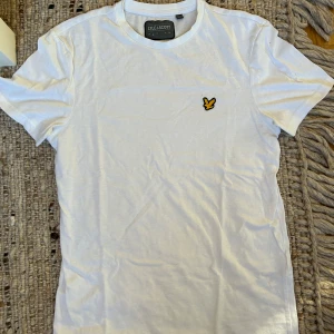 Vit t-shirt från Lyle & Scott - Vit t-shirt från Lyle & Scott med klassisk passform och korta ärmar. T-shirten har rund halsringning och den ikoniska gula örnloggan broderad på bröstet. Tillverkad i mjuk bomull, perfekt för en clean och stilren look.