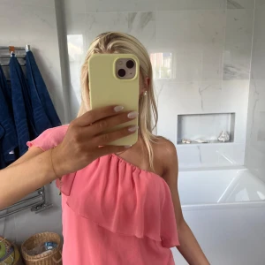 Rosa offshoulder blus med volang - Säljer en söt rosa blus i luftig polyester med volangdetalj upptill. Toppen kan bäras offshoulder eller oneshoulder och har en avslappnad passform. Perfekt för sommarens varma dagar och ger en fräsch och trendig look.