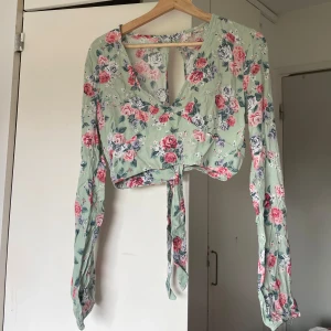 Blommig omlottopp - Superfin croppad omlottopp från H&M Divided i ljusgrönt med blommigt mönster i rosa, vitt och blått. Toppen har långa ärmar, v-ringning och knyt framtill. Perfekt för dig som gillar romantisk och trendig stil. Är L men passar M