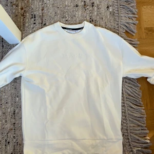 Vit sweatshirt från Calvin Klein - Säljer en stilren vit sweatshirt från Calvin Klein med broderad logga framtill. Tröjan har rund halsringning och långa ärmar. Perfekt för dig som gillar en clean och enkel look. Materialet är mjukt och skönt, troligtvis bomull.