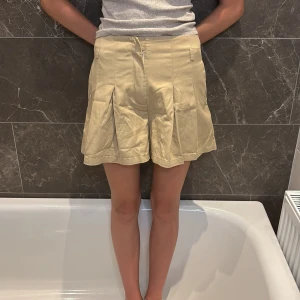 Beige shorts från Hian Tjen - Snygga beige shorts från Hian Tjen for Josephine Anni. De har hög midja, vida ben och snygga veck framtill som ger en cool och dressad look. Gjorda i ett mjukt bomullstyg med fickor och bälteshällor. Perfekta för sommarens alla dagar!