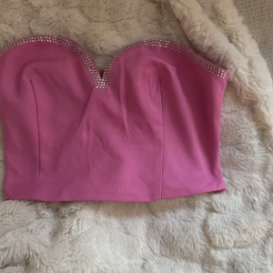 Rosa bandeau tubtopp från NA-KD - Supersnygg rosa bandeau tubtopp från NA-KD med glittriga stenar längs den hjärtformade urringningen. Toppen är axelbandslös och har smockad baksida för extra komfort. Perfekt för dig som vill sticka ut med en färgstark och trendig look.