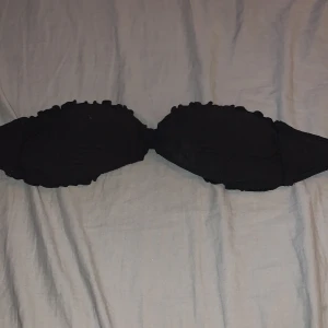 Svart bandeau bikiniöverdel med volang - Svart bandeau bikiniöverdel med volangdetaljer längs kanterna och knyt i ryggen. Bikinin har en lätt ribbad struktur och vadderade kupor för extra stöd. Perfekt för strandhäng och poolparty. Den är knappt använd och i storlek 34