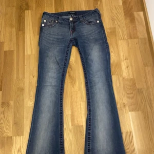 True Religion bootcut jeans blå - Snygga blå jeans från True Religion med bootcut passform och röda kontrastsömmar. Klassisk femficksmodell med ikonisk broderad logga på bakfickorna. Jeansen har en låg midja och är tillverkade i mjukt denimtyg. Säljer för att de tyvärr är för små på sista bilden ser ni min kille som provar jeansen 