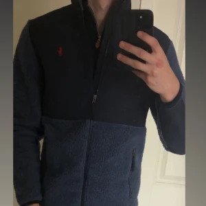 Blå teddyjacka från Polo Ralph Lauren - Säljer en blå fleece från Polo Ralph Lauren med dragkedja och hög krage. Jackan har mörkblå detaljer på bröst och ärmar, röd broderad logga och praktiska fickor med dragkedja. Perfekt för höst, vår och vinter dagar och riktigt snygg till jeans. Har bara använt 1 gång då den var för liten. Passar runt 180cm lång.