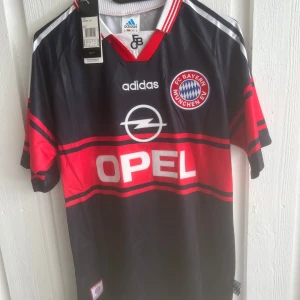 Bayern München Matthäus retrotröja Adidas - Klassisk Bayern München fotbollströja från Adidas med nummer 10 och Matthäus på ryggen. Svart bas med röda och vita detaljer, breda röda ränder och klubbmärke på bröstet. V-ringad krage med röd och vit rand, korta ärmar och tryckt Opel-logga framtill.