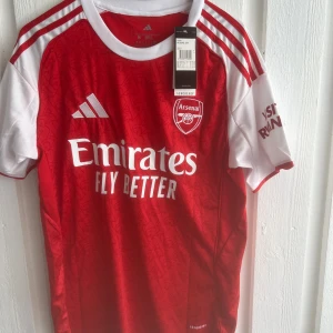 Arsenal Saka 7 matchtröja Adidas - Röd och vit Arsenal fotbollströja från Adidas med Saka 7 på ryggen. Klassisk design med vita axlar och ärmar, röda detaljer och klubbmärke på bröstet. Tillverkad i lätt och ventilerande material, perfekt för match eller träning.