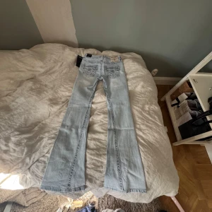 True Religion ljusa bootcut jeans - Säljer ett par ljusblå bootcut jeans från True Religion, modell Joey Flare Flap. Jeansen har snygga detaljerade bakfickor med lock och silverknappar, samt klassisk True Religion patch. Tillverkade i mjuk denim med lätt tvättad look. Dem är helt nya 
