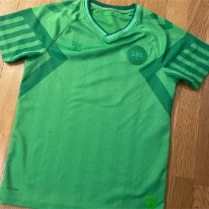 Danmark måliströja  152cm - Snygg grön fotbollströja från Hummel med DBU-logga på bröstet. Tröjan har V-ringning, korta ärmar med mörkgröna ränder och är tillverkad i ett lätt och ventilerande funktionsmaterial. Perfekt för träning eller match.