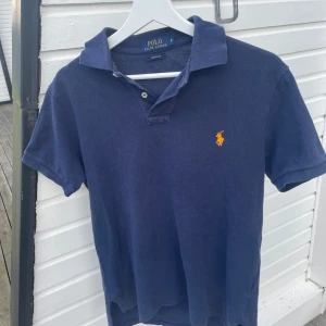 Marinblå pikétröja från Ralph Lauren - Klassisk marinblå pikétröja från Ralph Lauren i 100% bomull. Tröjan har korta ärmar, krage med två knappar och den ikoniska orange broderade loggan på bröstet. Perfekt för en stilren och avslappnad look. 