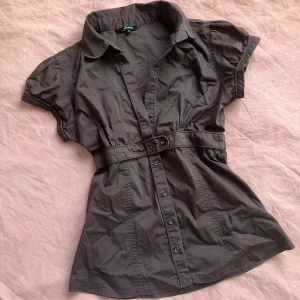 Brun kortärmad skjorta med bälte - Snygg brun skjorta med korta ärmar, markerad midja och tillhörande bälte. Skjortan har klassisk krage och knäppning framtill. Perfekt för dig som gillar en feminin och stilren look.