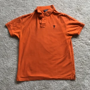 Orange pikétröja från Polo Ralph Lauren - Snygg orange pikétröja från Polo Ralph Lauren med klassisk krage, knappar framtill och den ikoniska broderade loggan på bröstet. Tröjan är i bomull och har korta ärmar, perfekt för en chill och sportig stil.