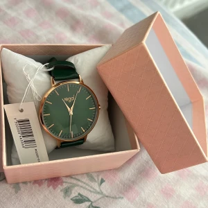 Grön klocka från Regal med gulddetaljer - Stilren klocka från Regal med mörkgrönt armband och matchande grön urtavla. Klockan är helt ny, aldrig använts dock finns det ingen box kvar till den. Därför ligger den i en annan låda🤗