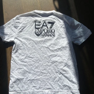 Vit EA7 Emporio Armani t-shirt - Vit t-shirt från EA7 Emporio Armani med svart logga tryckt stort på ryggen och en mindre patch på bröstet. Klassisk rund hals och korta ärmar. Perfekt för dig som gillar stilrena märkesplagg med tydlig branding.skick 10/10