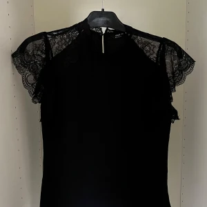 Svart spetsblus från ONLY XXS - Supersnygg svart blus från ONLY med transparenta spetsärmar och rund halsringning. Blusen har fina spetsdetaljer över axlarna och en liten knapp i nacken. 