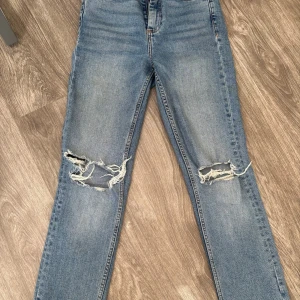 Blå slitna bootcut jeans med hål - Säljer ett par blå skinny jeans med slitna detaljer och stora hål över knäna. Jeansen har en klassisk femficksmodell och är i stretchigt denim som sitter tajt längs benen. Perfekta för en avslappnad och trendig look.