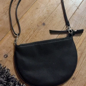 Svart axelväska i skinn med dragkedja - En stilren svart axelväska i äkta skinn med rundad form och silverfärgad dragkedja. Väskan har en smal axelrem och är perfekt för att bära det viktigaste. Enkel och klassisk design som passar till många outfits.