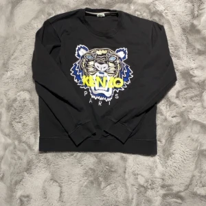Svart Kenzo sweatshirt med tiger - Svart sweatshirt från Kenzo med ikoniskt broderat tigerhuvud i blått, vitt, gult och grått. Stort Kenzo Paris-tryck framtill. Mjuk bomullskvalitet och klassisk passform med långa ärmar och ribbade muddar. Perfekt statement-plagg för dig som gillar streetstyle.