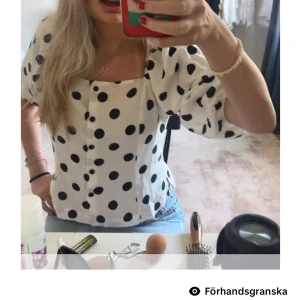 Vit prickig blus med puffärm - Supersöt vit blus med stora svarta prickar och puffiga korta ärmar. Blusen har fyrkantig ringning och knappar framtill, vilket ger en trendig och lekfull look. Perfekt att styla med jeans eller kjol för en cool outfit.