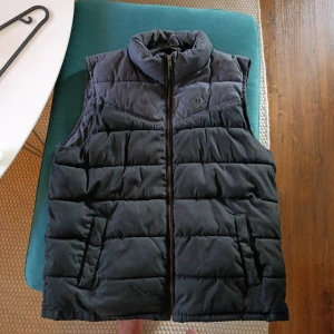 Svart dunväst från FSBN XL - Svart quiltad dunväst från FSBN i storlek XL. Västen har hög krage, två snedställda fickor och stängs med dragkedja framtill. Perfekt för lager-på-lager och har en stilren look med diskret logodetalj på bröstet.