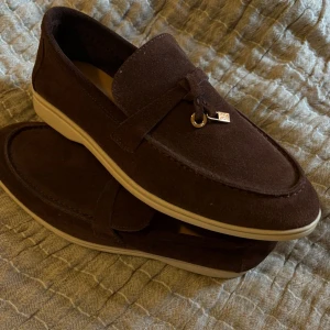 Bruna Loro Piana - Snygga loafers i mörkbrun mocka med beige sula och dekorativ knut framtill med liten metallberlock. Skorna har en klassisk, rund tå och är perfekta för dig som gillar stilrena och tidlösa skor med en twist. Storlek 39. Oandvända . Fick dom i present stora för mig . OBS inget kvitto 