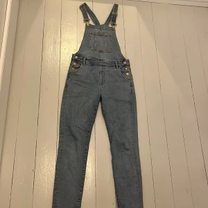 Ljusblå denim hängselbyxor MNG Kids - Snygga ljusblå denim hängselbyxor från MNG Kids med klassiska justerbara axelband, bröstficka och silverfärgade knappar i sidorna. Byxorna har raka ben och bakfickor, perfekta för en chill och trendig look.