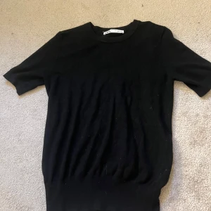 Svart stickad T-shirt från Zara - En svart, enkel och stilren stickad tröja från Zara med korta ärmar och rund hals. Perfekt basplagg som funkar till allt och är lätt att matcha. Mjuk och skön känsla, passar dig som gillar minimalistisk stil. Aldrig använd.