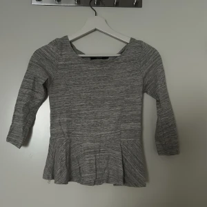 Grå peplum topp med trekvartsärm - Snygg grå peplum topp med trekvartsärm och rund halsringning. Toppen har ett diskret melerat mönster och är figursydd upptill med utsvängd nederdel. Perfekt att styla med jeans eller kjol för en trendig look.