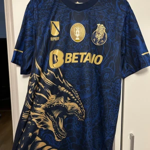 FC Porto blå guld fotbollströja drake - Snygg FC Porto fotbollströja i mörkblått med guldiga detaljer och stor drakdesign framtill. Tröjan har korta ärmar, tryckta loggor och sponsortryck i guld. Materialet är lätt och andas, perfekt för match eller träning. Unik look med coola mönster och klubbmärke på bröstet.