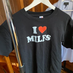 Svart t-shirt med I ❤️ MILFS-tryck - Svart t-shirt från Fruit of the Loom med vit text och rött hjärta på bröstet där det står 'I ❤️ MILFS'. Klassisk passform med rund hals och korta ärmar. Perfekt statement-plagg för dig som vill sticka ut och visa attityd.
