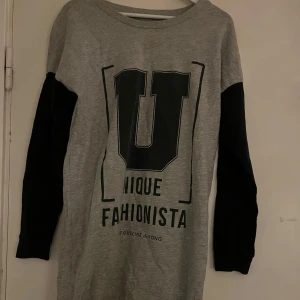 Grå långärmad tröja Jacqueline de Yong - Snygg grå långärmad tröja/ klänning med svarta ärmar och stort tryck framtill med texten 'Unique Fashionista'. Perfekt för en chill och trendig look. Mjuk bomullskänsla och relaxed passform.