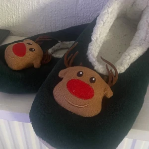 Mysiga tofflor med renmotiv - Supermjuka tofflor i mörkgrönt tyg med vit fluffig insida och en söt ren med röd nos framtill. Perfekta för att hålla fötterna varma under vintern och julen. Tofflorna har platt sula och är gjorda i syntetmaterial.