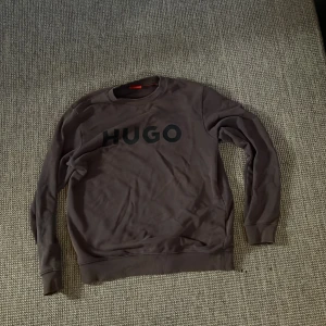 Brun sweatshirt från HUGO - Säljer en brun sweatshirt från HUGO med stor svart logga framtill. Tröjan har rund halsringning, långa ärmar och ribbade muddar vid ärmslut och nederkant. Perfekt för en chill och stilren look.