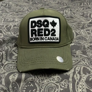 Olivgrön keps från Dsquared2 - Snygg olivgrön keps från Dsquared2 med broderad patch framtill där det står 'DSQ RED2 Born in Canada'. Justerbar baktill och broderad logga även där. Klassisk böjd skärm och cool streetkänsla. Perfekt för dig som vill sticka ut med en exklusiv accessoar.